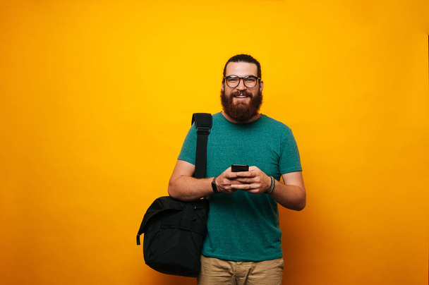 Jeune homme barbu portant des lunettes et des aéropodes regardant la caméra et souriant. Hipster portant des lunettes va travailler en utilisant le téléphone
. - Photo, image