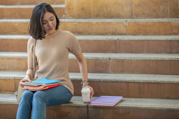 Beautiful Asian female college student holding her books and a cup of coffe sitting at stairs, people education learning high school program smart teenager concept - Photo, image