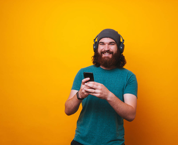 Cheerful bearded hipster man listen music at headphones and using mobile phone over yellow background - Photo, Image