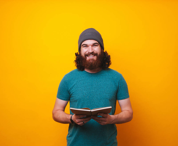 Happy cheerful bearded hipster man holding and reading book over yellow background - Foto, Imagem