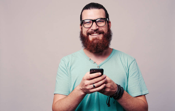 Ευτυχισμένος γενειοφόρος hipster άνθρωπος φορώντας γυαλιά και κρατώντας το κινητό τηλέφωνο σε γκρι φόντο - Φωτογραφία, εικόνα