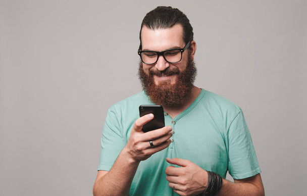 Молодой бородатый мужчина с телефоном на сером фоне
 - Фото, изображение