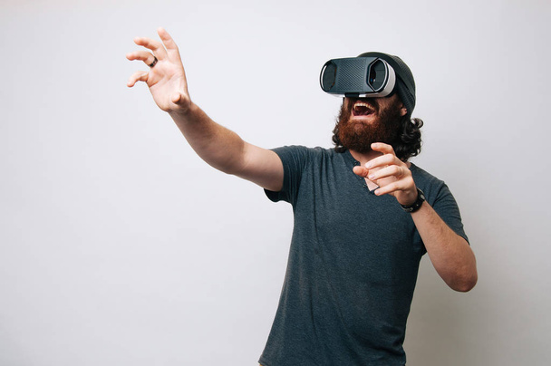 Веселый хипстер молодой бородатый мужчина смотрит в виртуальную реальность виртуальной реальности с современным цифровым гаджетом на белом фоне
. - Фото, изображение