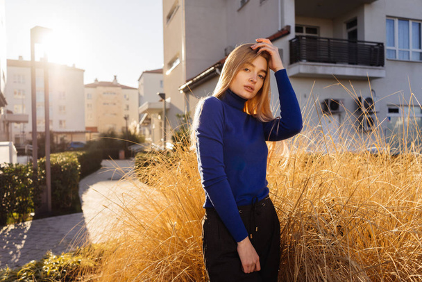 young blond model girl in blue sweater posing in sunshine outdoors rays - 写真・画像
