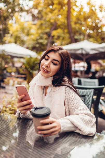 Beautiful young woman having coffee in outdoor cafe while using smartphone. Portrait of stylish smiling girl typing message in autumn park - Foto, afbeelding