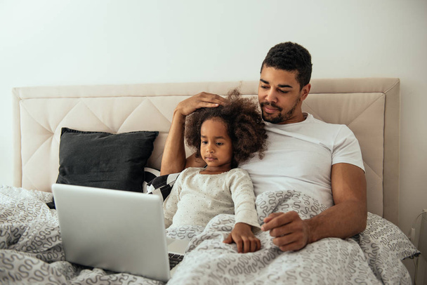 Portrait of an african american father and daughter lying in the bed and using laptop. - 写真・画像