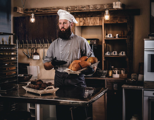 Guapo chef barbudo en uniforme mostrando bandeja de pan fresco en la cocina de panadería
. - Foto, Imagen