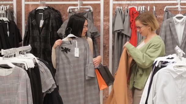 Young woman chooses a dress in a clothing store and consults with her friend. Trying on dresses in the mirror. Girl brunette is going to buy a dress and consults with blonde friend. - Footage, Video