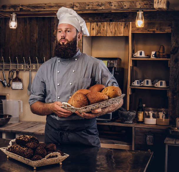 Guapo chef barbudo en uniforme mostrando bandeja de pan fresco en la cocina de panadería
. - Foto, imagen