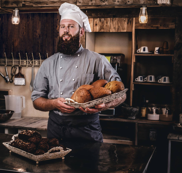 Bello chef barbuto in uniforme che mostra vassoio di pane fresco in cucina di panetteria
. - Foto, immagini