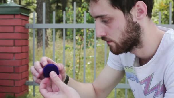 Good-looking guy opens small blue velvet box with wedding ring next to fence - Footage, Video
