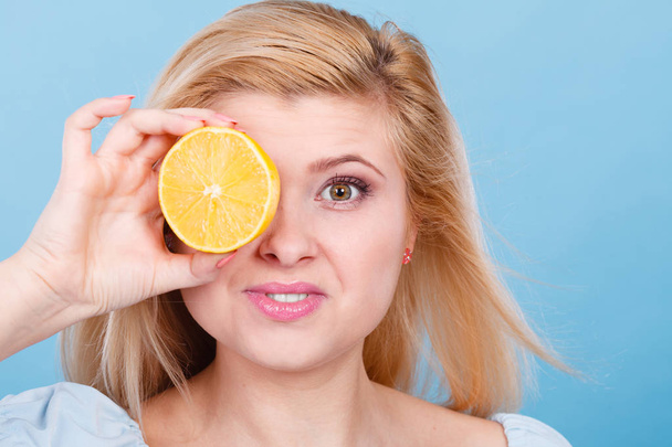 Woman teen girl holding half of yellow lemon citrus fruit in hand, covering her eye, on blue. Healthy diet nutrition. Happiness fun concept. - Photo, Image