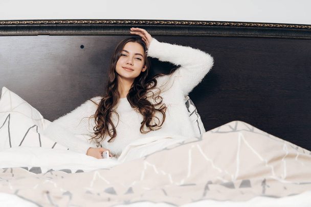 charming sleepy long-haired girl in white pajamas lies in bed, does not want to get up - Fotografie, Obrázek