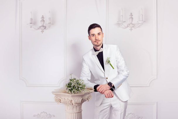 Trendy groom in white suit and black shoes standing with bridal bouquet on small column near looking at camera posing. Half body portrait - Zdjęcie, obraz