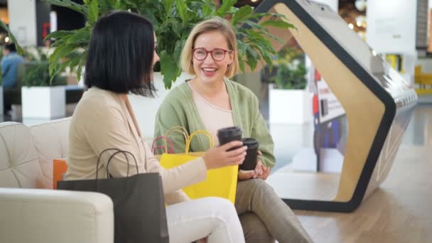 Two young women drinking coffee, talking and laughing at cafe in the mall after shopping. People, friendship, conversation and togetherness concept - Footage, Video
