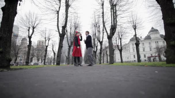 Ensayo de hombre en chaqueta de terciopelo y mujer en falda roja bailando juguetona y alegremente tango por sendero de hormigón en zona de parque en temporada de invierno, rodeado de árboles oscuros sin hojas
. - Metraje, vídeo