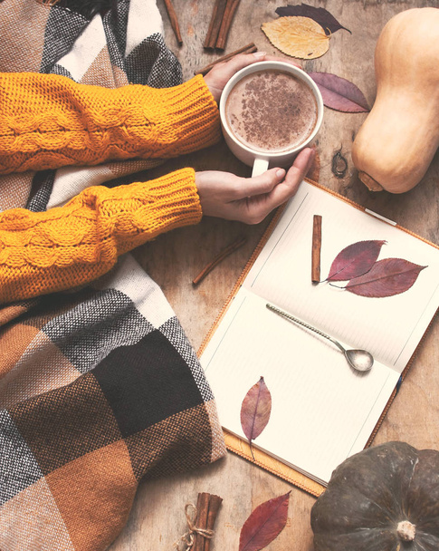 Υπαίθρου ξύλινο υπόβαθρο με κολοκύθα και φθινοπωρινά φύλλα, τα χέρια της γυναίκας με ένα φλιτζάνι καφέ latte κολοκύθα ή κακάο, το σημειωματάριο, καρό σε ένα κλουβί, το top view ήπια επίδραση - Φωτογραφία, εικόνα
