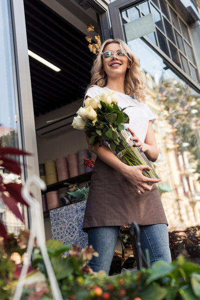 vista angolo basso di bella fiorista felice uscire dal negozio di fiori con rose in vaso
 - Foto, immagini