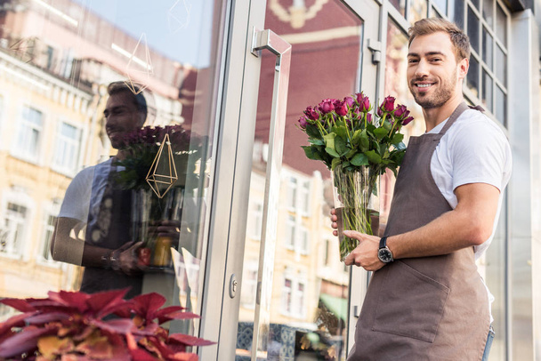 низкий угол обзора красивого флориста, держащего стеклянную банку с бордовыми розами возле цветочного магазина и смотрящего в камеру
 - Фото, изображение