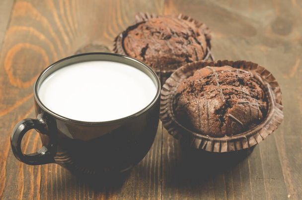 two chocolate muffins and milk on a wooden background/two chocolate muffin in a white plate and a black mug with milk. Selective focus - Foto, Bild