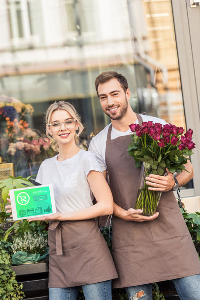 Fleuristes souriants tenant tablette avec appareil commercial et roses bordeaux près de la boutique de fleurs
 - Photo, image