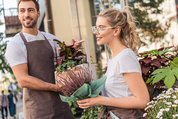 smiling florists holding potted plants near flower shop and looking away - Foto, Imagem