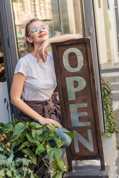 attractive smiling flower shop owner leaning on open sign - Foto, Imagen