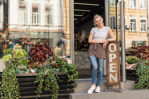 smiling attractive florist standing on stairs of flower shop and leaning on open sign - Фото, зображення