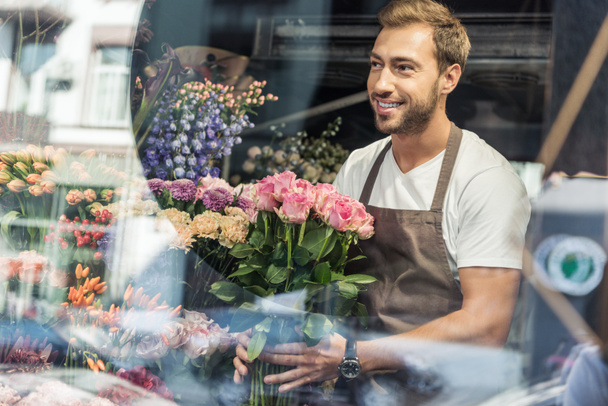 view through window of handsome florist holding bouquet of pink roses in flower shop and looking away - Foto, Imagen