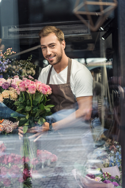 view through window of handsome florist holding bouquet of pink roses in flower shop and looking at camera - Фото, изображение