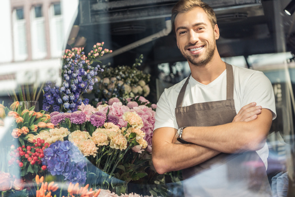 view through window of handsome florist standing with crossed arms in flower shop and looking at camera - Fotoğraf, Görsel