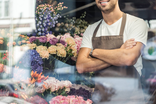 imagen recortada de florista sonriente de pie con los brazos cruzados en la florería
 - Foto, Imagen