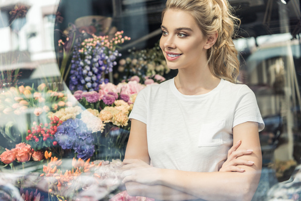 view through window of beautiful florist standing with crossed arms in flower shop and looking away - Photo, Image