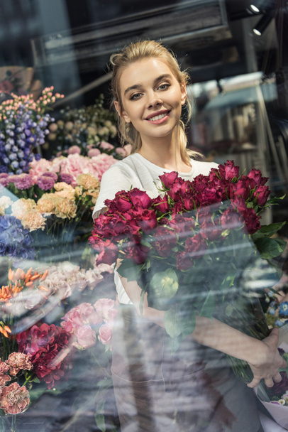 view through window of attractive florist holding bouquet of burgundy roses in flower shop and looking at camera - Photo, Image