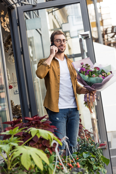 χαμογελώντας όμορφος άνδρας πηγαίνει από το κατάστημα λουλουδιών με μπουκέτο και μιλάμε με smartphone - Φωτογραφία, εικόνα