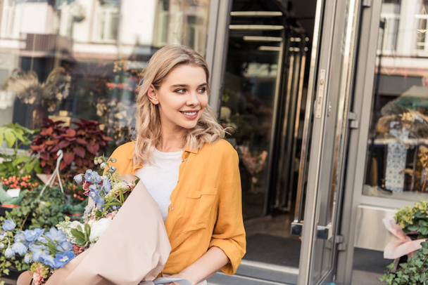 portrait of attractive smiling girl going out from flower shop with wrapped bouquet and looking away - Φωτογραφία, εικόνα