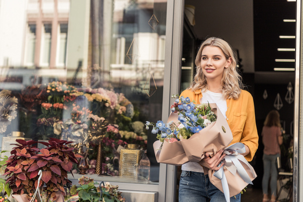 beautiful woman going out from flower shop with wrapped bouquet and looking away - Foto, afbeelding