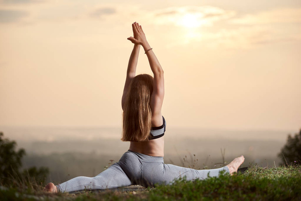 Back view of long-haired athletic woman with raised arms doing yoga exercises sitting in green grass on bright pink foggy sky at sunset copy space background. Fitness and healthy lifestyle concept. - Фото, изображение