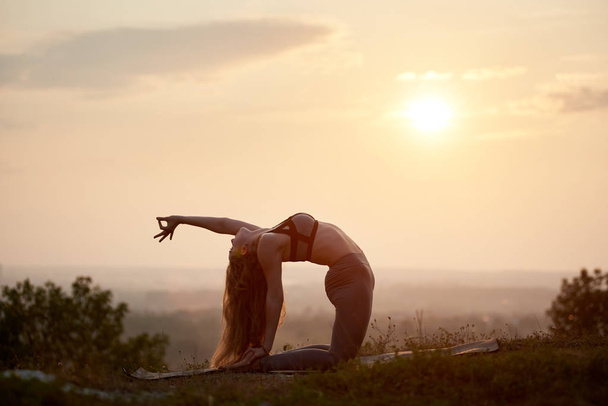 Sylwetka atrakcyjny smukły sprawny elastyczne długowłosy młoda kobieta robi skomplikowane ćwiczenia gimnastyczne jogi na świeżym powietrzu na trawiastą wzgórzu na tle jasne różowe niebo zamglone o zachodzie słońca. - Zdjęcie, obraz