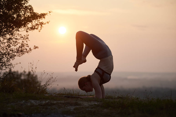 Silhouet van sterke flexibele blote voeten meisje in opleiding outfit doen moeilijk ingewikkelde yoga oefeningen buiten op achtergrond van licht roze zomer avondlucht. Fitness en beauty concept. - Foto, afbeelding