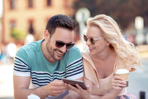 sonriente joven pareja sentada en la mesa en la cafetería mientras mira el teléfono inteligente y beber café
 - Foto, Imagen