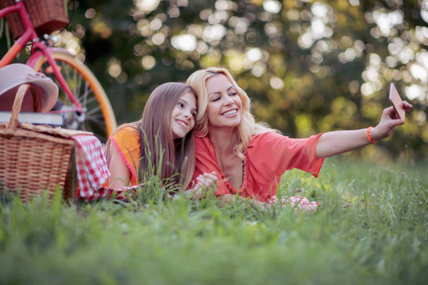 beautiful mother and daughter having picnic in park and taking selfie on smartphone in daylight - Photo, image