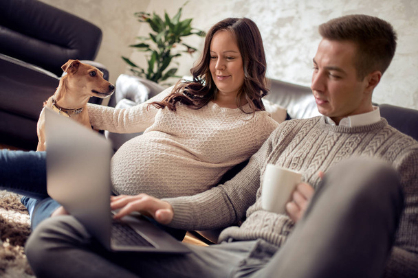 Young family, pregnant woman and her husband with dog sitting on sofa . - Photo, Image