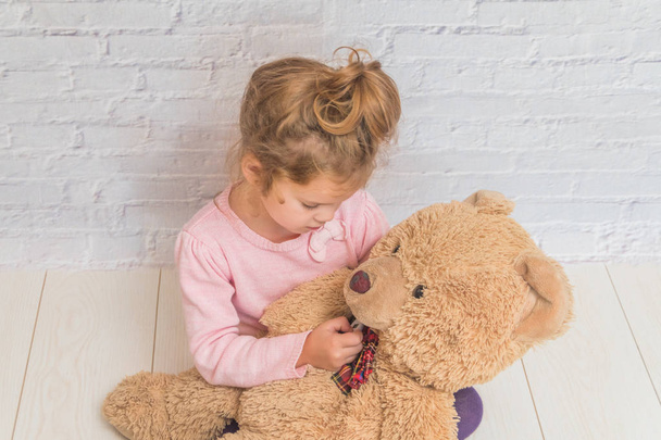 the girl, child measures the temperature of a toy bear, against a white brick wall - Foto, imagen