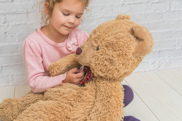 the girl, child measures the temperature of a toy bear, against a white brick wall - Foto, Bild