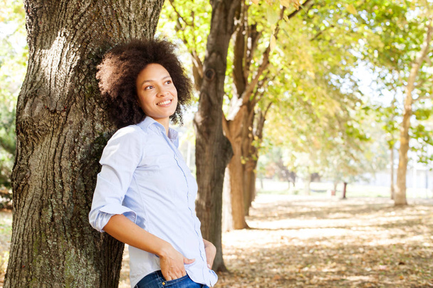 Outdoor portrait of happy beautiful african american woman in nature, female in blue shirts enjoy summer day,  leaning on tree in park, looking at the side   - Valokuva, kuva