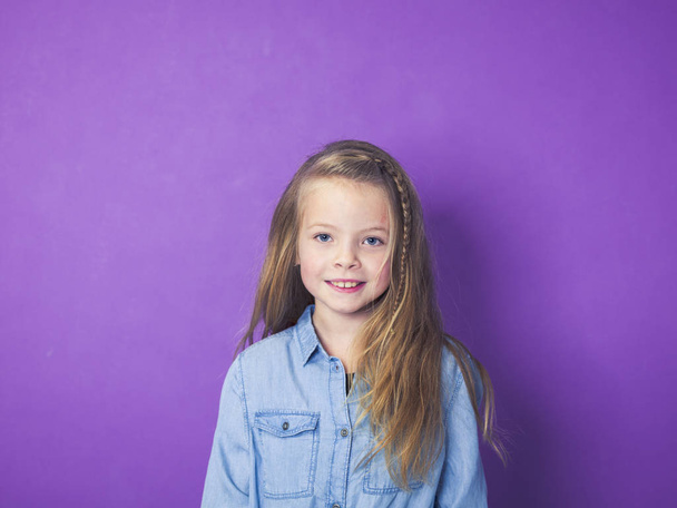 portrait of cute little girl wearing blue denim shirt in front of violet background in studio - Photo, Image