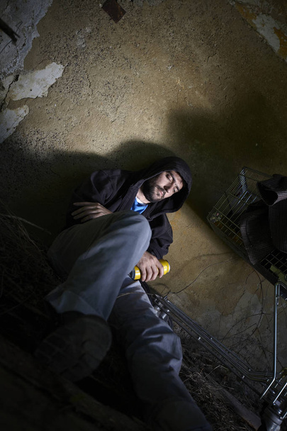 bearded homeless man in dirty hoodie sitting on floor with beer can in hand at abandoned house  - Photo, Image
