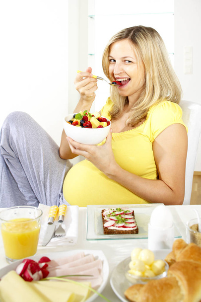 happy pregnant woman sitting on chair and eating fruit salad from bowl while sitting near table with dishes  at home - Zdjęcie, obraz