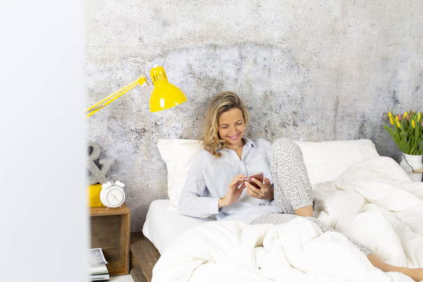 young blonde woman typing message on smartphone while lying in bed  - Foto, Imagen